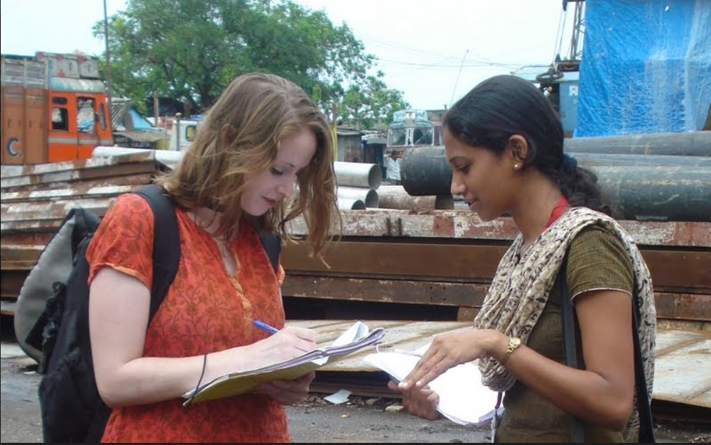 Heather Lanthorn is conducting an interview. She is writing on papers on top of a notebook with a blue pen. Heather is a white woman wearing an orange shirt and a backpack speaking with an interviewee on the street.